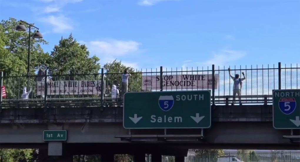 A group of people on an overpass hanging black and white signs that read “Stop White Genocide” and “Make White Babies.”