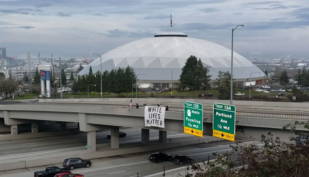 A group of people hang a white banner with black text over an overpass. The banner reads “White Lives Matter.”