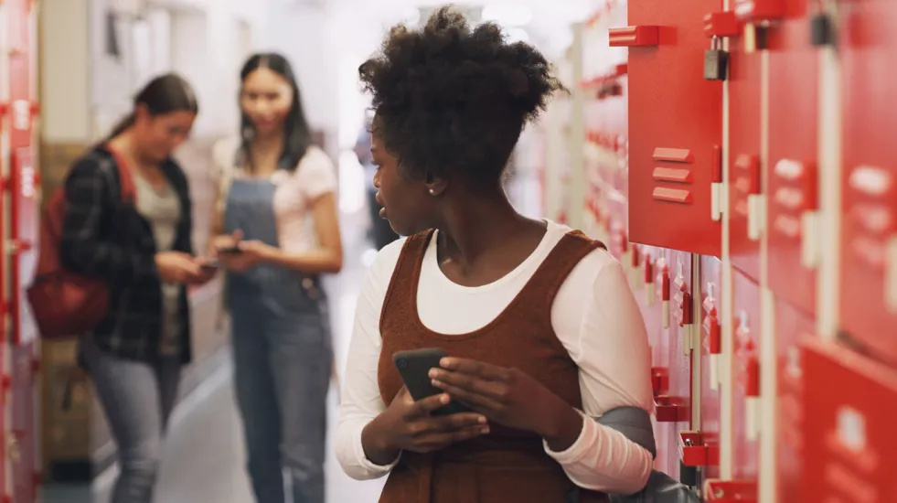 Teenage girl standing next to her school locker using a smartphone and being bullied