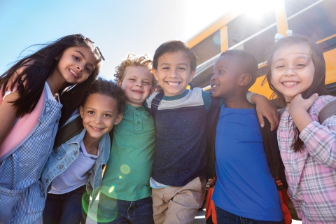 A group of diverse children with arms around each other standing in front of a school bus smiling